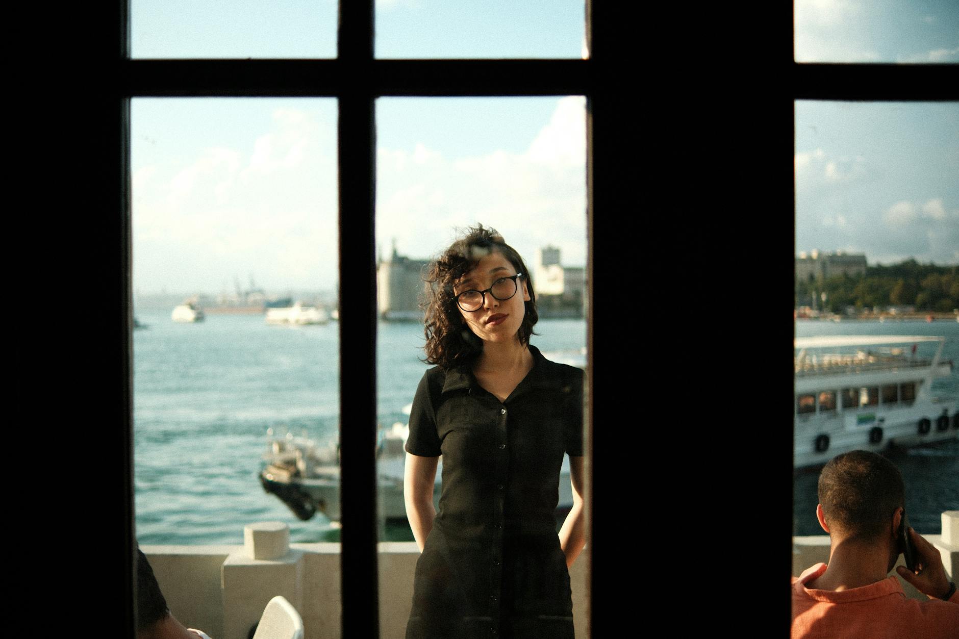 woman leaning against a window with a view of sea and ships behind her